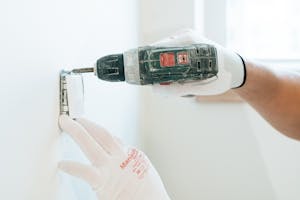 Gloved hands using a drill to install a power outlet on a wall, focusing on precision and safety.